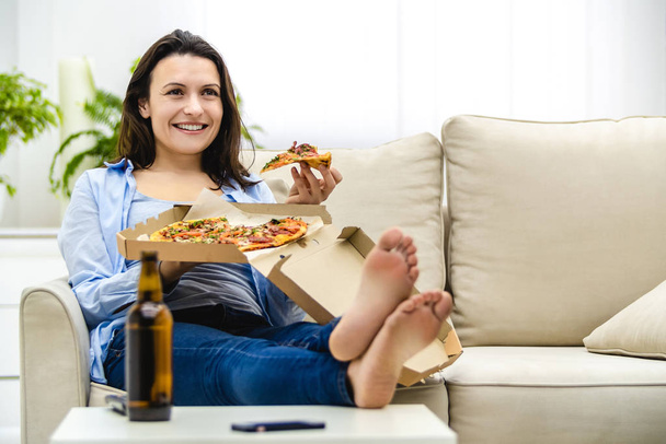 Beautiful and happy woman is eating pizza. She is smiling sincerely, while sitting on the sofa. Copy space. - Foto, Bild