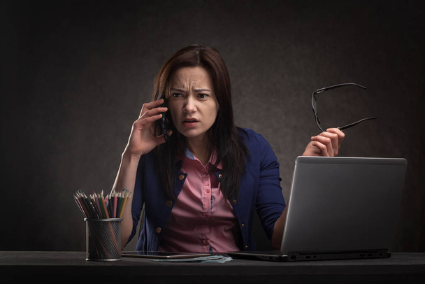 Успешная деловая женщина за столом на тёмном фоне разговаривает по телефону
. - Фото, изображение