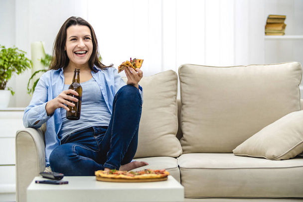 Atraktivní žena pije studené pivo a jí pizzu, zatímco se dívá na zápas na TV. Je nesmírně šťastná. Kopírovat prostor. - Fotografie, Obrázek