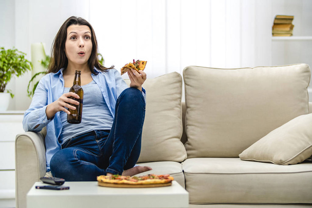 Attractive and interested woman is shocked, watching television. Amazed woman is sitting on the lisht sofa and eating snacks. - 写真・画像