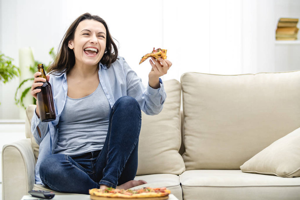 Attractive woman is drinking cold beer and eating pizza, while watching the game on TV. - Fotografie, Obrázek