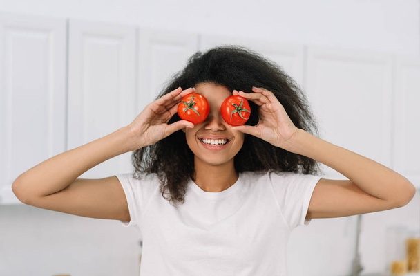 Šťastné emoce Afroameričanka drží červená rajčata blízko očí, stojí v kuchyni, baví se. Zdravý životní styl, vegetariánská strava. Atraktivní emocionální dívka vaření čerstvý salát  - Fotografie, Obrázek