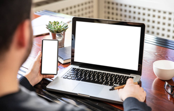 Portátil y smartphone con pantallas blancas en blanco, fondo interior de oficina
 - Foto, Imagen