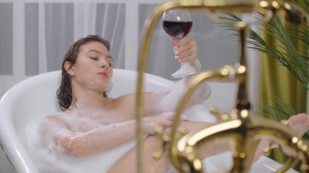 schöne junge brünette Frau genießt angenehmes Bad mit Schaum, liegt mit geschlossenen Augen und hält ein Glas Wein. - Filmmaterial, Video