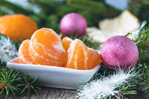 Fette di mandarino giallo maturo si trovano in una ciotola accanto ai rami verdi di un albero di Natale e giocattoli di Natale.
. - Foto, immagini
