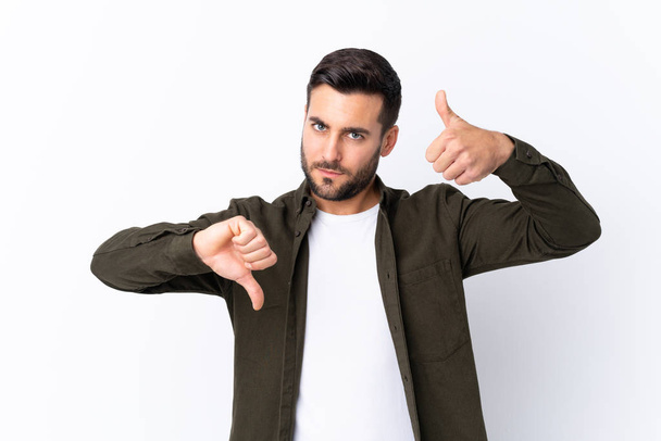 Young handsome man with beard over isolated white background making good-bad sign. Undecided between yes or not - Φωτογραφία, εικόνα