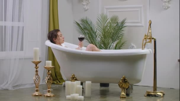 Zijaanzicht van een vermoeid jong krullend meisje liggend, ontspannend in het bad binnen en luisterend naar de muziek vormen een draadloze luidspreker naast haar en een glas rode wijn op de voorgrond. - Video