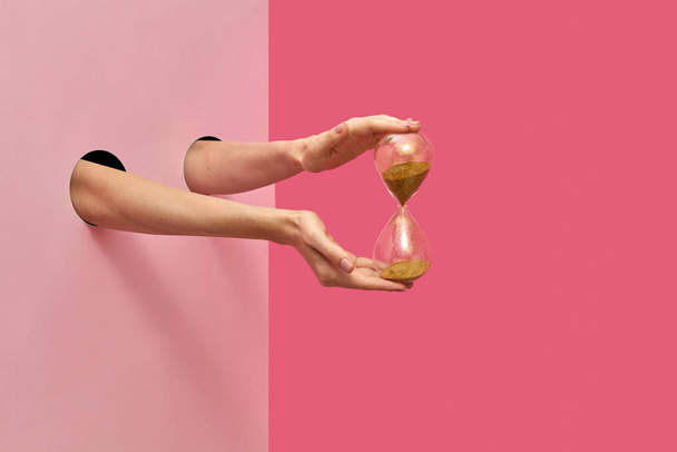 Vintage sandglass in a female's hands from two holes in the wall on a duotone pink background, copy space. - Φωτογραφία, εικόνα