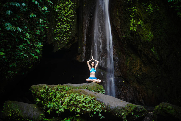 Young Caucasian woman meditating, practicing yoga at waterfall. Hands raising up in namaste mudra. Leke Leke waterfall, Bali, Indonesia.  - Photo, Image