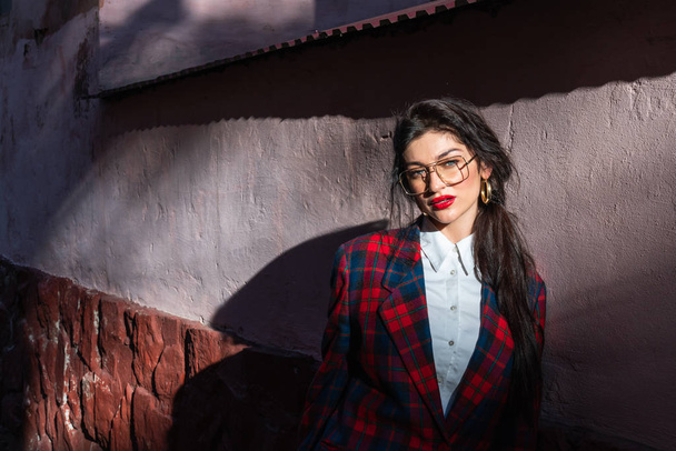 A brooding attractive girl with black hair, glasses and red lips, dressed in a plaid jacket and white shirt, stands near the wall in the sun and looks at the camera - Photo, Image