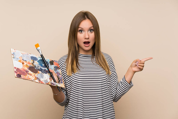 Teenager dívka drží paletu přes izolované pozadí překvapené a ukazuje prstem na stranu - Fotografie, Obrázek