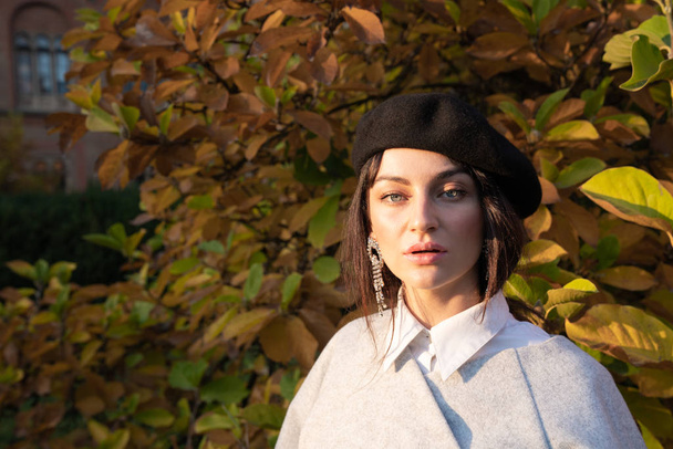 Close up portrait of the beautiful brunette girl with grey eyes in black beret looking at the camera, yellow leaves on the background - Fotó, kép