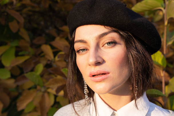 Close up portrait of the beautiful brunette girl with grey eyes in black beret looking at the camera, yellow leaves on the background - Photo, Image