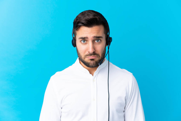Telemarketer άνθρωπος που εργάζονται με ένα ακουστικό πάνω από απομονωμένο μπλε φόντο λυπηρό - Φωτογραφία, εικόνα