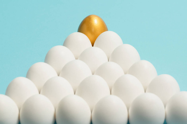 Concept van individualiteit, exclusiviteit, betere keuze. Een gouden ei onder witte eieren op blauwe achtergrond. - Foto, afbeelding