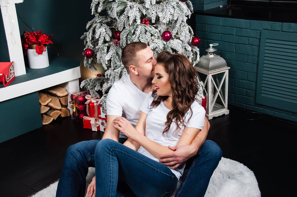 Szerető pár, férfi és nő, ölelkeznek és nézik egymást az újévi belső térben közel karácsonyfa és kandalló - Fotó, kép