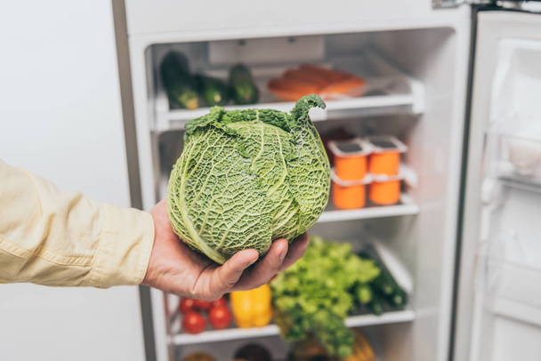 cropped view of man holding cabbage near open fridge with fresh food on shelves - Photo, Image