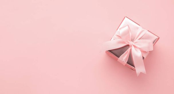 Caixa festiva em embalagem de ouro com um arco de cetim em um fundo rosa pastel. Layout de estilo plano. Conceito de férias
. - Foto, Imagem
