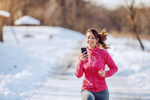 Uśmiechnięta biała brunetka w odzieży sportowej i ze słuchawkami w uszach patrzącymi na smartfon podczas biegania w naturze. Zimą. Koncepcja sprawności na zewnątrz. - Zdjęcie, obraz