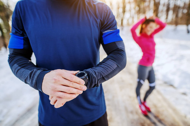 Fit sporty caucasian man in sportswear checking out on smart watch heart rate after running. In background is his female friend tying hair. Wintertime. Outdoor fitness concept. - Photo, Image