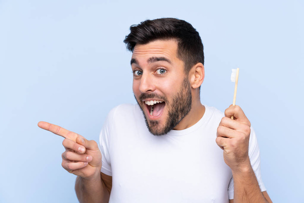 Young handsome man with beard brushing his teeth over isolated background surprised and pointing finger to the side - Foto, afbeelding