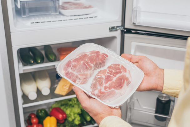 cropped view of man holding frozen meat near open fridge full of food - Foto, Imagem