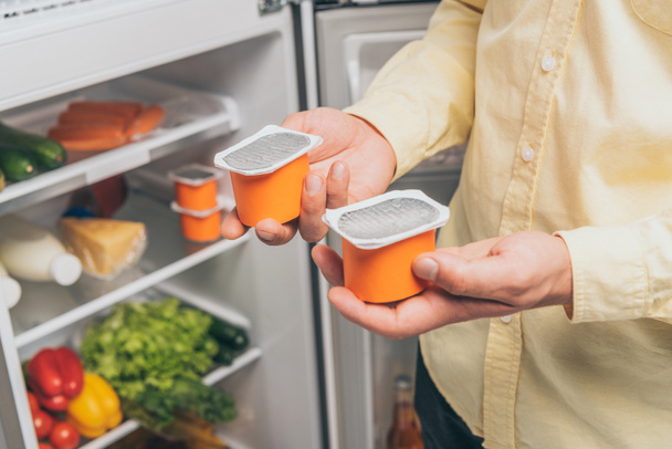cropped view of man holding yogurt near open fridge full of food - Zdjęcie, obraz
