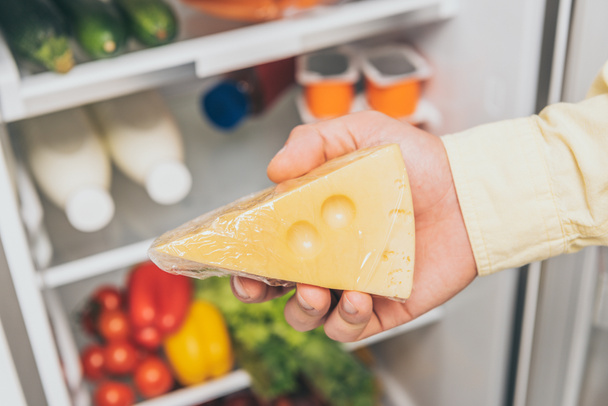 обрізаний вид людини, що тримає сир біля відкритого холодильника, повного їжі
 - Фото, зображення