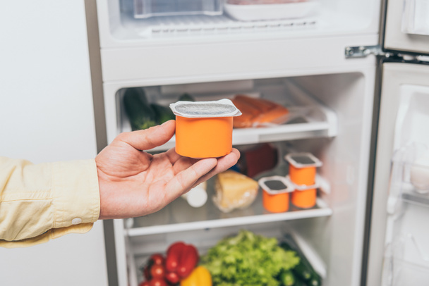abgeschnittene Ansicht eines Mannes, der Joghurt in der Nähe eines offenen Kühlschranks voller Lebensmittel hält - Foto, Bild