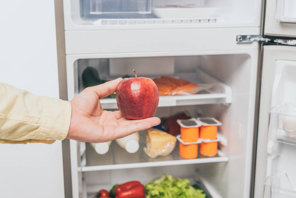 cropped view of man holding red apple near open fridge full of food - Zdjęcie, obraz