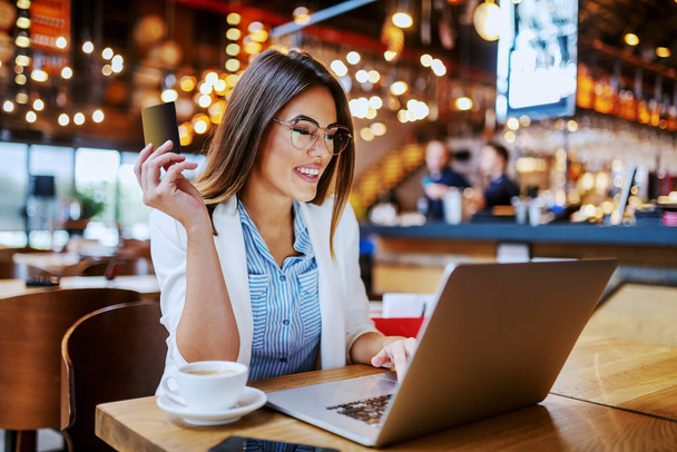 Superbe brune caucasienne souriante à la mode assise dans un café, tenant une carte de crédit et utilisant un ordinateur portable pour la recherche sur Internet. Après avoir magasiné dans les magasins, elle a décidé d'acheter plus d'articles en ligne
. - Photo, image