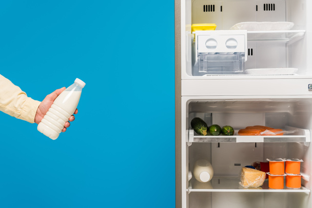 vista recortada del hombre sosteniendo la leche cerca de nevera abierta y congelador con alimentos frescos en estantes aislados en azul
 - Foto, Imagen