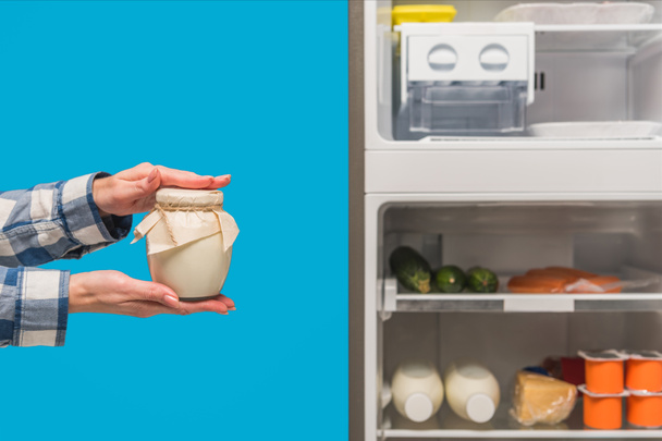 abgeschnittene Ansicht einer Frau mit saurer Sahne in der Nähe von offenem Kühlschrank und Tiefkühltruhe mit frischen Lebensmitteln in blau isolierten Regalen - Foto, Bild