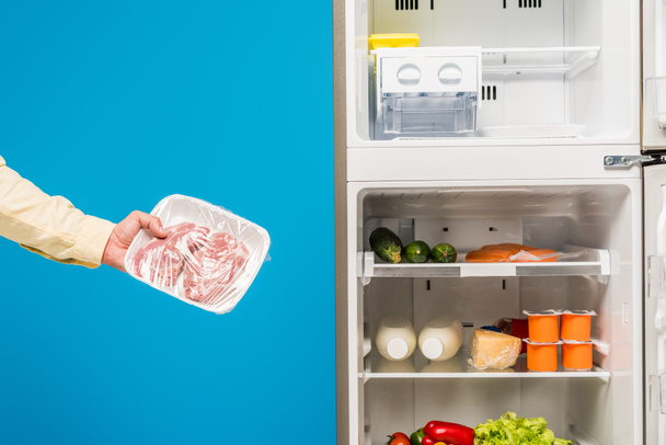 cropped view of man holding frozen meat near open fridge and freezer with fresh food on shelves isolated on blue - Foto, imagen