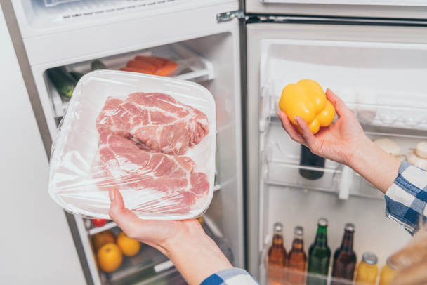 cropped view of woman holding frozen meat and bell pepper near open fridge with fresh food on shelves - Photo, Image