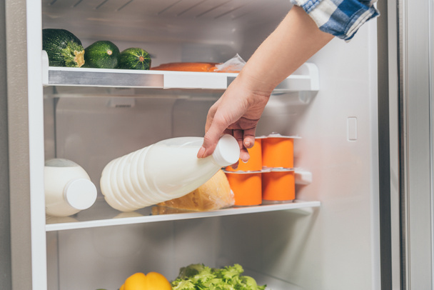 cropped view of woman taking bottle of milk out from fridge with fresh food on shelves - Photo, Image