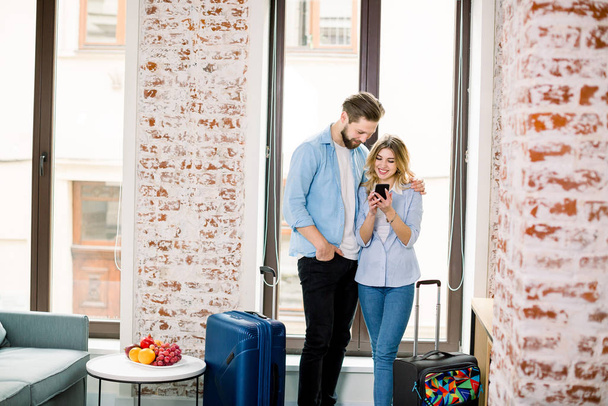 Happy couple looking for the photos in a smart phone standing over the window background in modern hotel room with brick walls - Foto, imagen