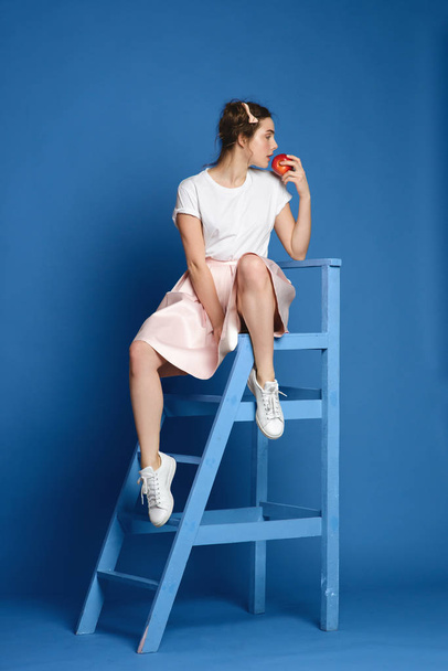 Красивая молодая женщина сидит на лестнице на классическом синем фоне стены
. - Фото, изображение