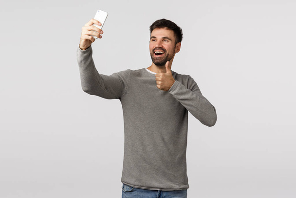 Komea ystävällinen ja lähtevä valkoihoinen mies harmaassa puserossa puhuu ystävän kanssa verkossa, käyttäen videopuhelun älypuhelinsovellusta, joka osoittaa näytön onnittelevaksi ystäväksi hyviä uutisia, hymyilevä hyväksyntä
 - Valokuva, kuva
