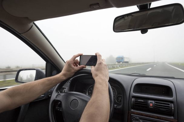 Uso de smartphone en el coche. Hombre usando el teléfono mientras conduce en la niebla en la carretera
. - Foto, imagen