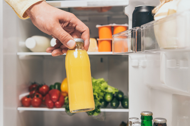 abgeschnittene Ansicht eines Mannes mit einer Flasche Saft in der Nähe eines offenen Kühlschranks mit frischen Lebensmitteln in den Regalen - Foto, Bild