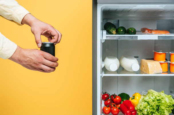 cropped view of man opening  can with soda near open fridge with fresh food on shelves Isolated On yellow - Foto, Imagen
