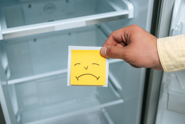 περικοπή άποψη του ανθρώπου κρατώντας λυπημένος smiley κοντά άδειο ανοιχτό ψυγείο - Φωτογραφία, εικόνα