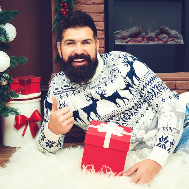 Bearded guy lying at fireplace at Christmas. Gifts and greetings for Christmas. Happy New Year. Fashion man in christmas sweater at home - Фото, изображение