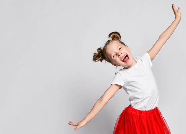 Singing laughing kid girl in fatin skirt and t-shirt is posing holding her hands diagonal spread on white - Фото, изображение