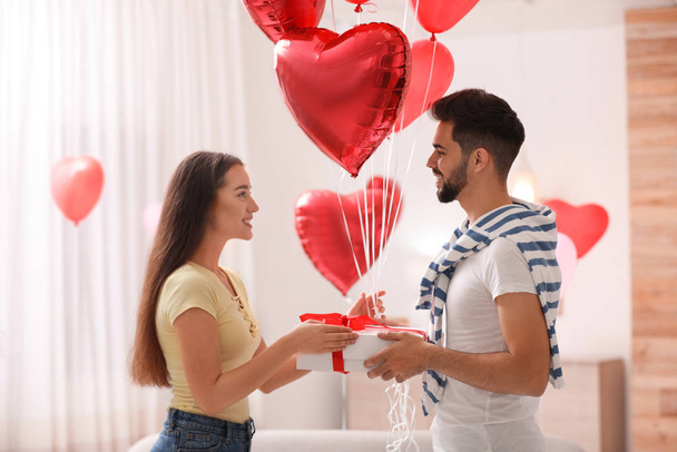 Lovely young couple with gift box and heart shaped  balloons in living room. Valentine's day celebration - Valokuva, kuva