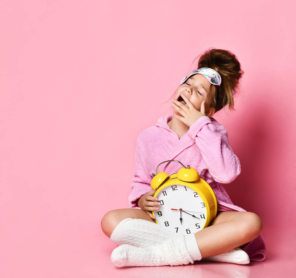 Yawning teenage girl holding an alarm clock while sitting in a housecoat on the floor. - 写真・画像