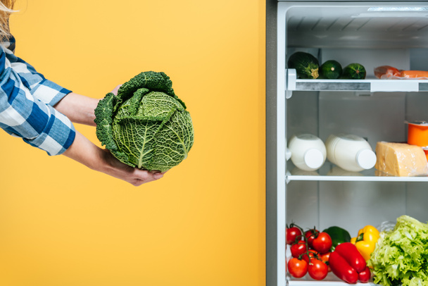cropped view of woman holding cabbage near open fridge with fresh food on shelves Isolated On yellow - Foto, Bild