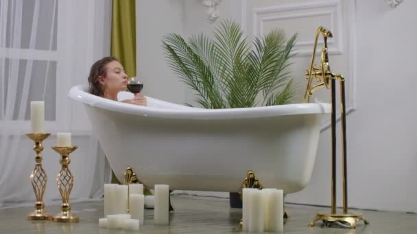 leuke jonge vrouw in wit shirt met wijnglas nemen van een bad. Sensueel meisje geniet in de badkamer met. Vrouw in wit shirt drinkt rode wijn uit. - Video