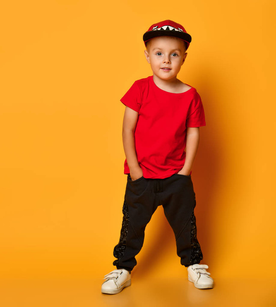 Young guy boy in a red T-shirt and dark pants, white sneakers and a funny cap posing on a free copy space on a yellow background - Foto, Imagem